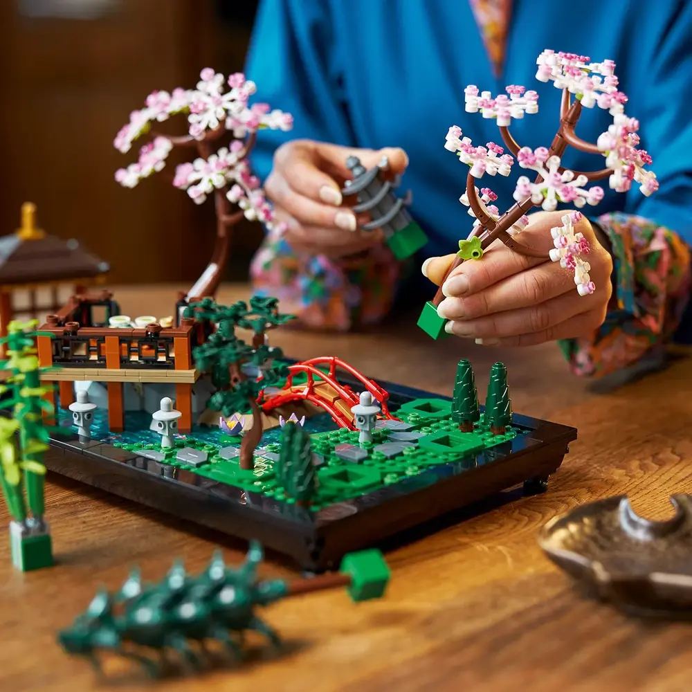 Mãos de uma pessoa montando uma das cerejeiras em flor do set LEGO Jardim Tranquilo 10315, com o jardim parcialmente construído ao fundo.